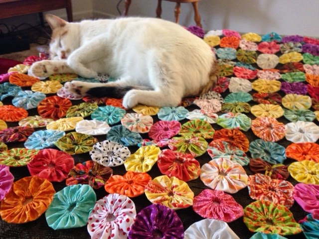 A white cat with orange spots is sleeping curled up on a colorful, handmade quilt crafted from fabric yo-yo sewing projects using various patterned circles.