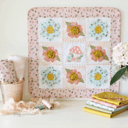 A pastel-colored quilt with floral, mushroom patterns, and fabric yo-yos is displayed upright. Beside it are folded fabrics, sewing threads, and a potted hydrangea, creating a cozy, creative space for fabric crafts.