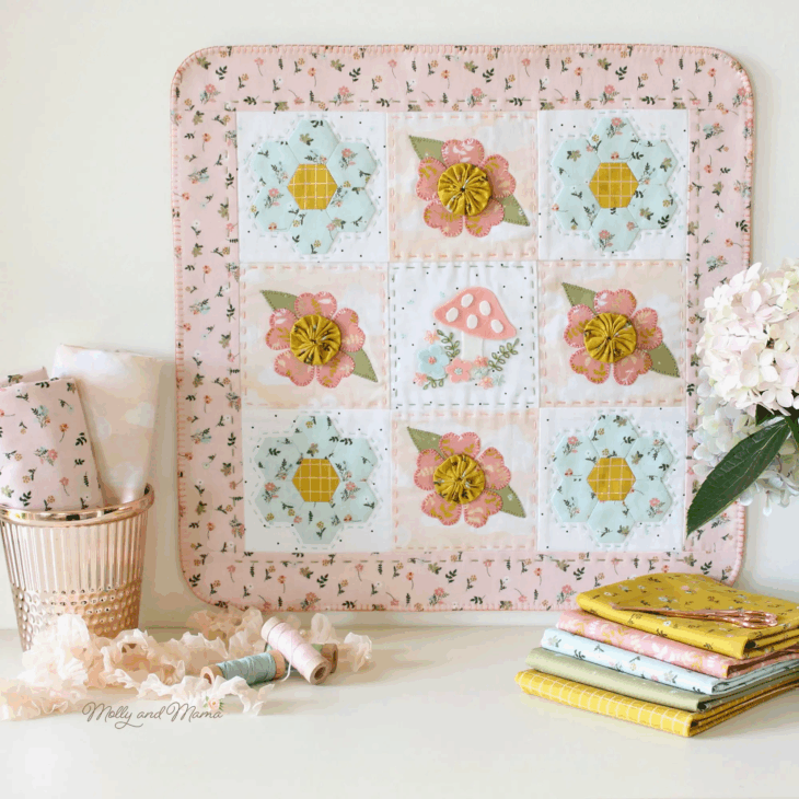 A pastel-colored quilt with floral, mushroom patterns, and fabric yo-yos is displayed upright. Beside it are folded fabrics, sewing threads, and a potted hydrangea, creating a cozy, creative space for fabric crafts.
