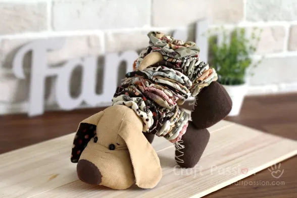 A handmade fabric dog figurine crafted from stacked, colorful fabric yo-yos stands on a wooden surface. The background features a blurred "Family" sign and a small green plant, perfect inspiration for fabric crafts enthusiasts.