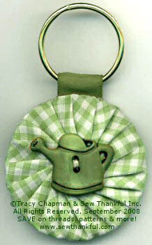 A round keychain made of green and white gingham fabric features a fabric yo-yo with a small green watering can decoration in the center&mdash;a charming accessory for lovers of fabric crafts. The keychain includes a sturdy metal ring at the top.