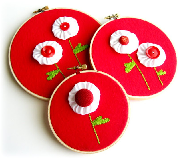 Three embroidery hoops with red fabric feature white fabric yo-yo flowers with red button centers and green embroidered stems and leaves, arranged in overlapping circles on a white background&mdash;an inspiring idea for fabric crafts and sewing projects.