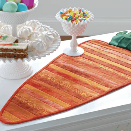 A carrot-shaped table runner with orange and green stripes—perfect for spring table runner patterns—sits on a white table, next to a tray of carrot cake, jellybeans, and pastel Easter eggs with white meringues.