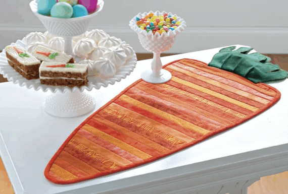 A carrot-shaped table runner with orange and green stripes&mdash;perfect for spring table runner patterns&mdash;sits on a white table, next to a tray of carrot cake, jellybeans, and pastel Easter eggs with white meringues.