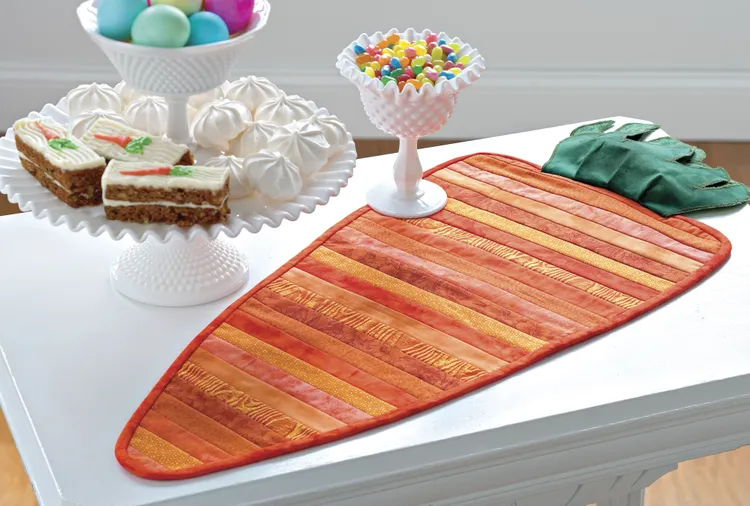 Easy Spring Table Runner Patterns You Can Sew in a Day 11 A carrot-shaped table runner with orange and green stripes—perfect for spring table runner patterns—sits on a white table, next to a tray of carrot cake, jellybeans, and pastel Easter eggs with white meringues.