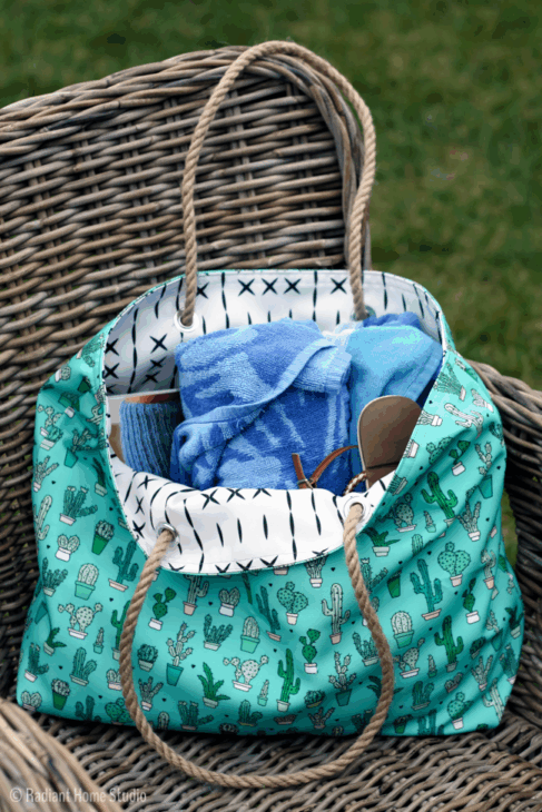 Sew Your Own Beach Bag or Tote with These Free Patterns 10 A green tote bag with cactus prints sits on a wicker chair, filled with blue towels and sunglasses. This stylish beach bag features rope handles, a white lining with black cross patterns, and inspiration for free patterns. Grass is visible in the background.