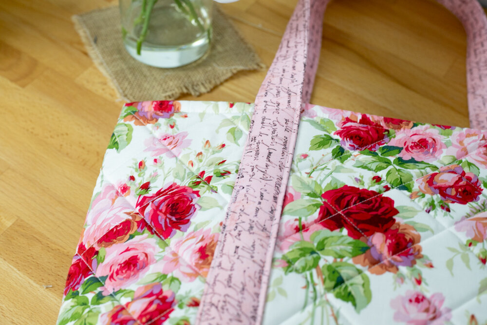 Sew Your Own Beach Bag or Tote with These Free Patterns 12 A quilted tote bag with pink handles and a floral pattern featuring red, pink, and white roses, rests on a wooden surface near a vase of flowers—perfect inspiration for those looking for stylish tote bag patterns.