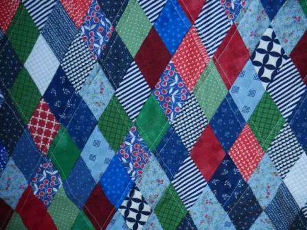 A colorful patchwork quilt with diamond-shaped pieces in blue, green, red, and white features various patterns&mdash;stripes, florals, polka dots, and checks&mdash;inspired by classic diamond quilt patterns.
