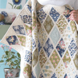 A person holds a handmade patchwork quilt featuring diamond quilt patterns with floral and patterned fabric. The background includes a blue wall, a chair with cushions, a watering can, and a stuffed toy.