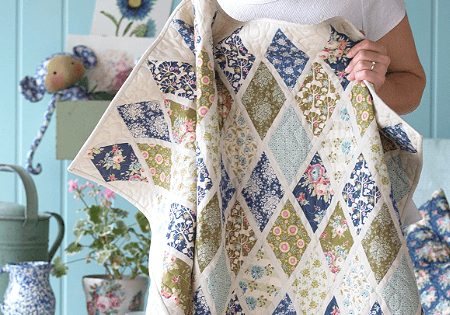 A person holds a handmade patchwork quilt featuring diamond quilt patterns with floral and patterned fabric. The background includes a blue wall, a chair with cushions, a watering can, and a stuffed toy.