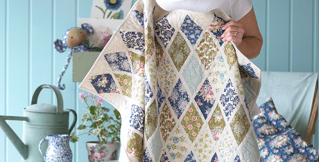 A person holds a handmade patchwork quilt featuring diamond quilt patterns with floral and patterned fabric. The background includes a blue wall, a chair with cushions, a watering can, and a stuffed toy.