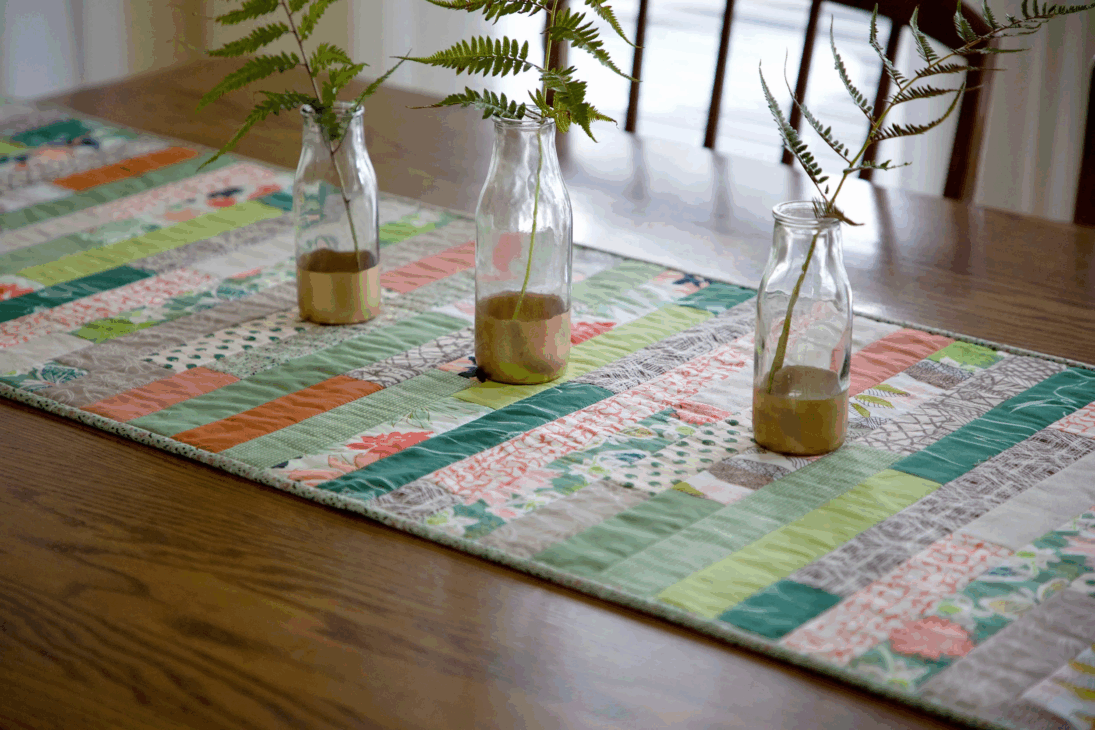 Easy Spring Table Runner Patterns You Can Sew in a Day 12 A wooden dining table with a colorful patchwork spring table runner pattern, topped with three glass bottles used as vases, each holding a single fern branch—an easy sewing project you could sew in a day.