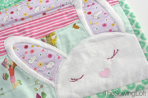 Easy Spring Table Runner Patterns You Can Sew in a Day 14 A close-up of a colorful quilt featuring an appliqué of a bunny face with closed eyes and a pink heart-shaped nose, surrounded by pastel fabrics with bunny and book patterns—perfect inspiration for Spring Table Runner Patterns to sew in a day.