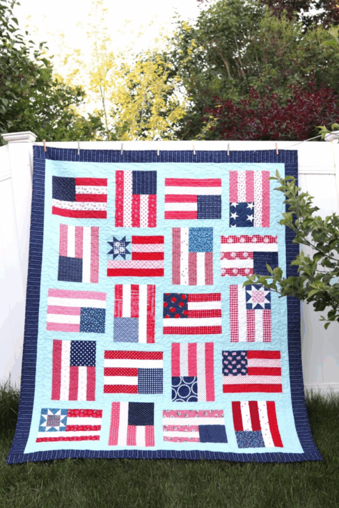 10 Patriotic Flag Projects to Sew Before the 4th of July 10 A colorful quilt featuring various stylized American flags—perfect for patriotic flag projects or to sew for the 4th of July—hangs on a white fence with greenery and trees in the background.