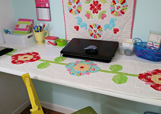 Easy Spring Table Runner Patterns You Can Sew in a Day 6 A neat craft workspace features a floral quilted table runner—perfect inspiration for easy sewing or sew in a day Spring table runner patterns—a closed laptop, colorful notebooks, a yellow mug with pens, and organized supplies beneath a bright wall quilt.