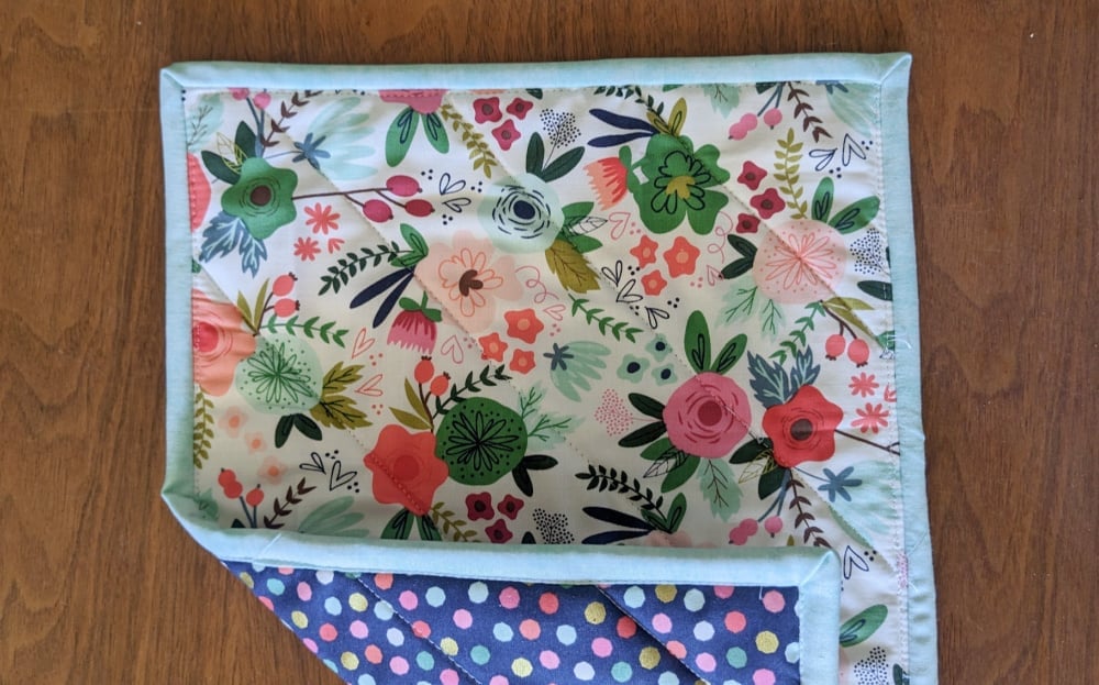 A small rectangular fabric hot pad with a floral pattern on one side and a multicolored polka dot pattern on the other, lying on a wooden surface. The hot pad has light blue edging.