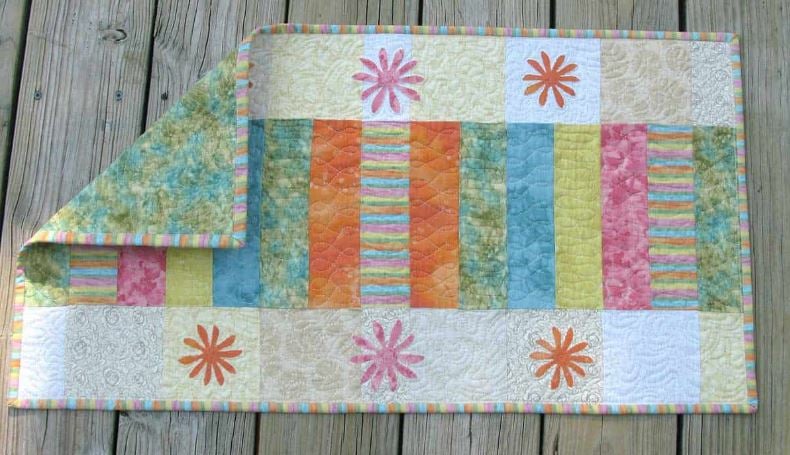 Easy Spring Table Runner Patterns You Can Sew in a Day 13 A colorful quilt with vertical striped panels in orange, teal, yellow, and pink, bordered by floral appliqués and a pastel striped edge—an easy sew table runner perfect for quick sewing projects—displayed on a wooden surface with one corner folded over.