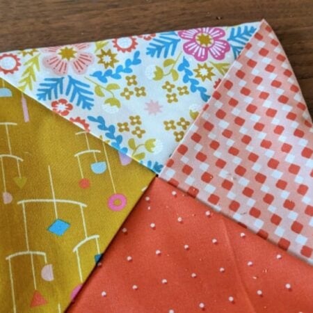 A hot pad fabric square folded to show four triangles, each with a different colorful pattern: yellow with mobiles, white with flowers, red with white dots, and red with a checkered design, resting on a wooden surface.