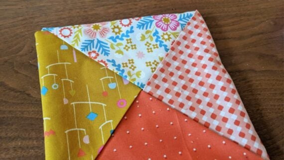 A hot pad fabric square folded to show four triangles, each with a different colorful pattern: yellow with mobiles, white with flowers, red with white dots, and red with a checkered design, resting on a wooden surface.