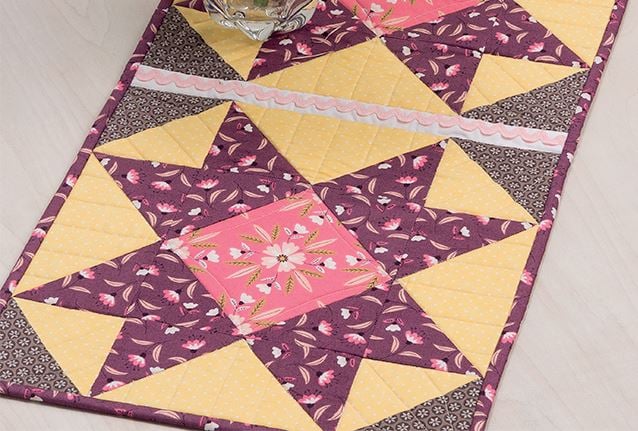 Easy Spring Table Runner Patterns You Can Sew in a Day 15 A quilted table runner featuring a star pattern with yellow, pink, and purple floral fabrics, accented with rickrack trim—an Easy Table Runner perfect for adding charm to your space. Displayed on a light-colored surface.