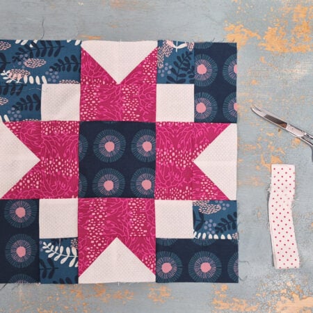 A colorful Quilt Block Swap piece with pink, white, and dark blue patterned fabrics is laid flat on a distressed gray surface. To the right are silver scissors and a folded strip of white fabric with red dots.