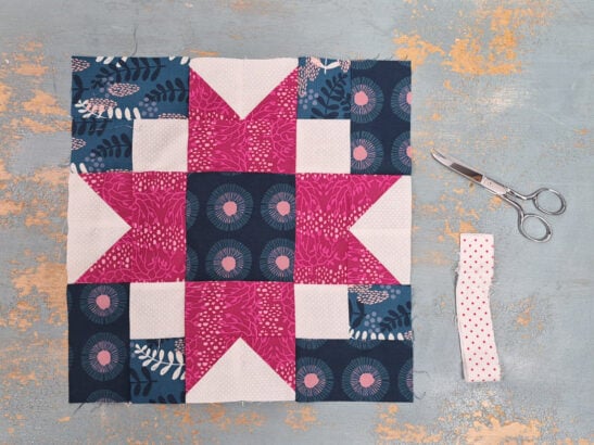 A colorful Quilt Block Swap piece with pink, white, and dark blue patterned fabrics is laid flat on a distressed gray surface. To the right are silver scissors and a folded strip of white fabric with red dots.