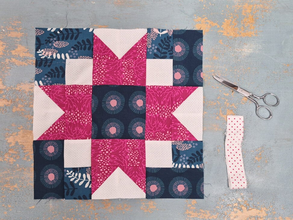 Christmas Quilt Block Swap - sign up is open! 1 A colorful Quilt Block Swap piece with pink, white, and dark blue patterned fabrics is laid flat on a distressed gray surface. To the right are silver scissors and a folded strip of white fabric with red dots.