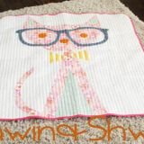 A charming cat quilt featuring a pink cat with large blue glasses, stitched on a white background and bordered with pink piping, is displayed on a beige carpet. The text "Shwin & Shwin" appears at the bottom.