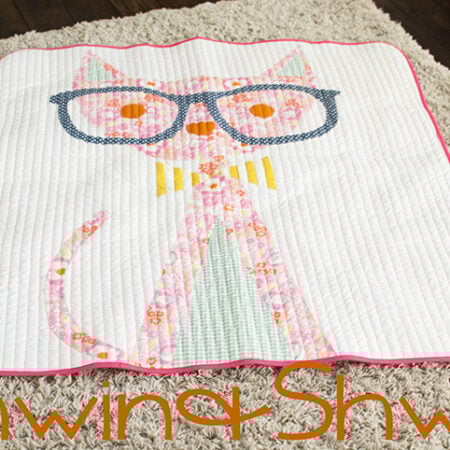 A charming cat quilt featuring a pink cat with large blue glasses, stitched on a white background and bordered with pink piping, is displayed on a beige carpet. The text "Shwin & Shwin" appears at the bottom.