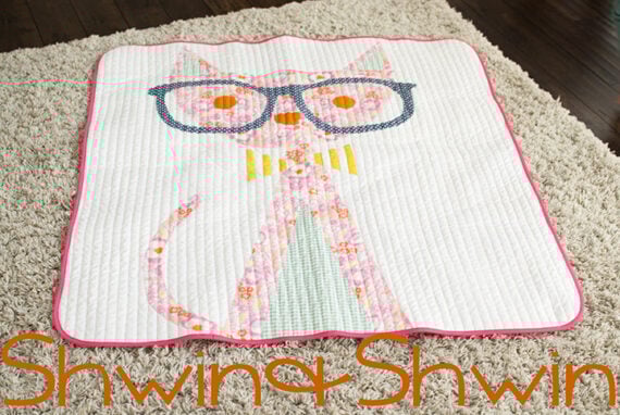 A charming cat quilt featuring a pink cat with large blue glasses, stitched on a white background and bordered with pink piping, is displayed on a beige carpet. The text "Shwin & Shwin" appears at the bottom.