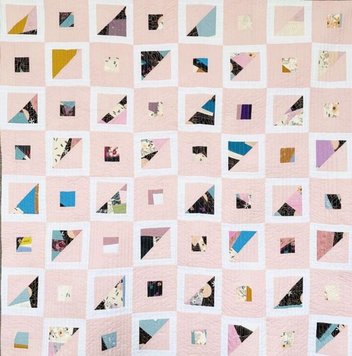 25 Must Sew Scrappy Strip Quilt Patterns 19 A handmade quilt with a grid of pastel pink squares, each displaying abstract geometric patterns in assorted colors and prints, inspired by scrappy strip quilt patterns. The playful mix of triangles and irregular shapes gives it a fresh, modern look.