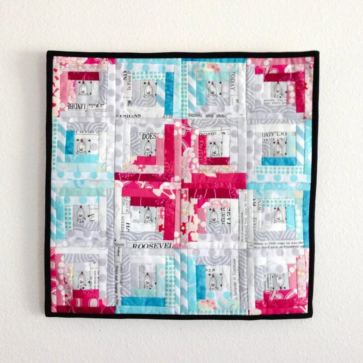 25 Must Sew Scrappy Strip Quilt Patterns 12 A colorful patchwork quilt inspired by scrappy strip quilt patterns features pink, blue, white, and gray fabric squares with various patterns and text, arranged in a geometric design and bordered with black edging, hanging on a white wall.