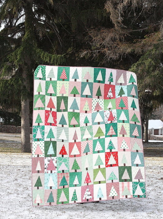 25 Must Sew Scrappy Strip Quilt Patterns 23 A colorful scrappy strip quilt featuring a repeating pattern of stylized Christmas trees in various shades of green, red, pink, and white is displayed outdoors before tall evergreens on a lightly snow-dusted lawn.