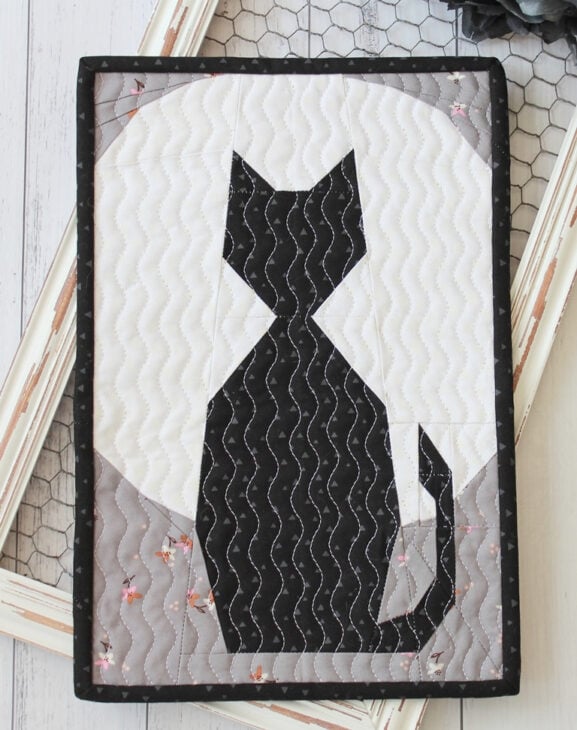 Quilted cat quilt wall hanging featuring a black cat silhouette against a gray and white circular background, accented by wavy stitching patterns. The cozy quilt rests on a rustic white wooden surface.