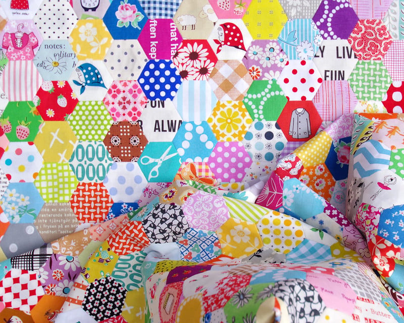 25 Must Sew Scrappy Strip Quilt Patterns 22 A colorful assortment of hexagon-shaped fabric pieces with various patterns and prints, arranged in a patchwork style inspired by scrappy strip quilt patterns, featuring polka dots, florals, and text fabrics in the foreground.