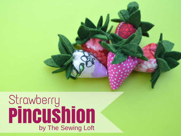 A group of fabric strawberry pincushions with green felt leaves sits on a bright green surface. Text reads "Strawberry Pincushion by The Sewing Loft" in pink and maroon, perfect inspiration if you love to sew pincushions or collect free pincushion patterns.