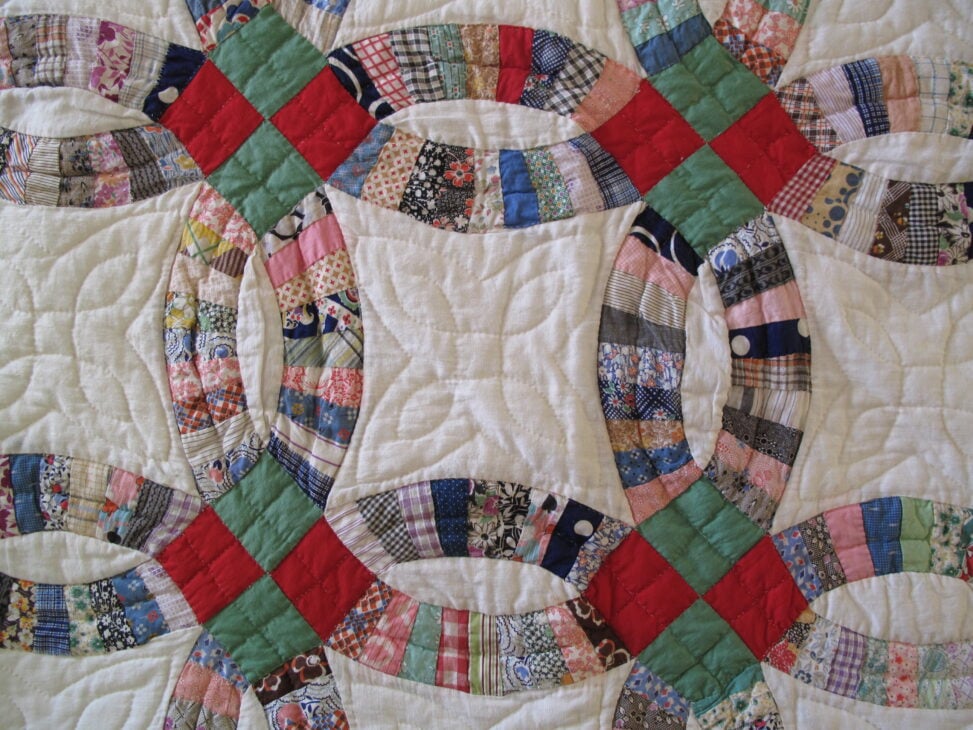 25 Must Sew Scrappy Strip Quilt Patterns 24 A close-up of a patchwork quilt showcases interlocking ring designs, crafted with scrappy strip quilt patterns in vibrant red, green, and multicolored fabrics on a white background. Visible stitching adds texture to the cheerful composition.