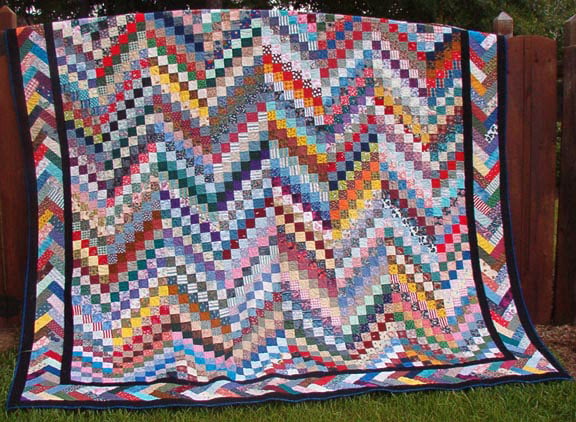 25 Must Sew Scrappy Strip Quilt Patterns 10 A colorful patchwork quilt, inspired by scrappy strip quilt patterns, features hundreds of small fabric squares in a zigzag arrangement, draped outdoors over a wooden fence with grass visible at the bottom.