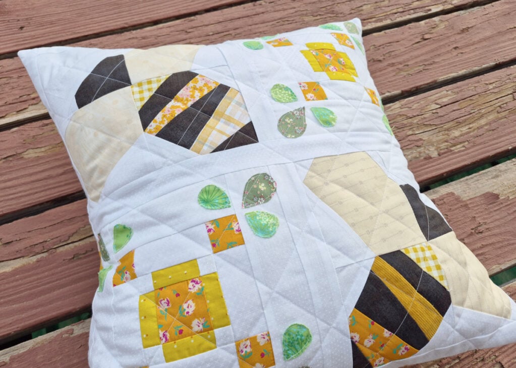 A quilted pillow with yellow, black, and patterned fabric blocks and green leaf shapes is placed on a weathered wooden surface. The pillow features a geometric, patchwork design.