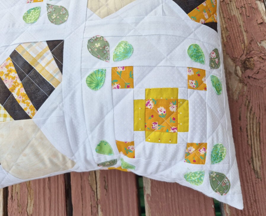 A quilted Bud pillow with a patchwork design featuring yellow floral squares and green leaf shapes on a white background, placed on a weathered wooden surface.
