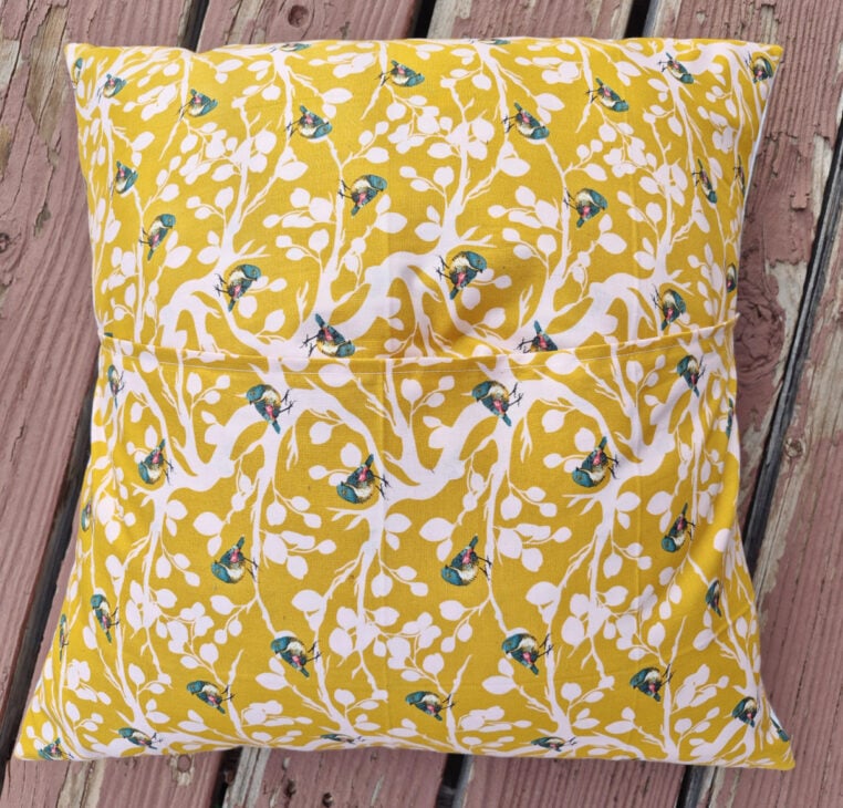 A square Bee & Bud pillow with a yellow cover featuring a white leafy pattern and colorful birds, resting on weathered wooden boards.