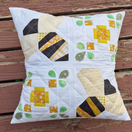 A quilted pillow with a bees and honeycomb pattern in yellow, black, and white fabrics, accented with green leaves, resting on a weathered wooden surface.