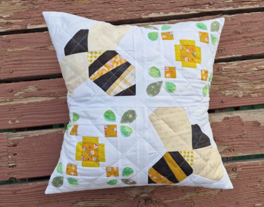 A quilted pillow with a bees and honeycomb pattern in yellow, black, and white fabrics, accented with green leaves, resting on a weathered wooden surface.