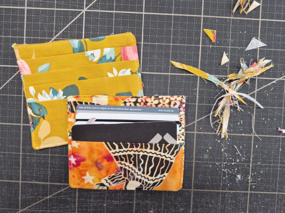 A handmade fabric Sew Card Wallet with colorful patterns sits on a cutting mat next to loose fabric scraps. Several cards are placed inside the wallet, and more pieces of yellow floral fabric are nearby&mdash;an easy to sew and functional project.