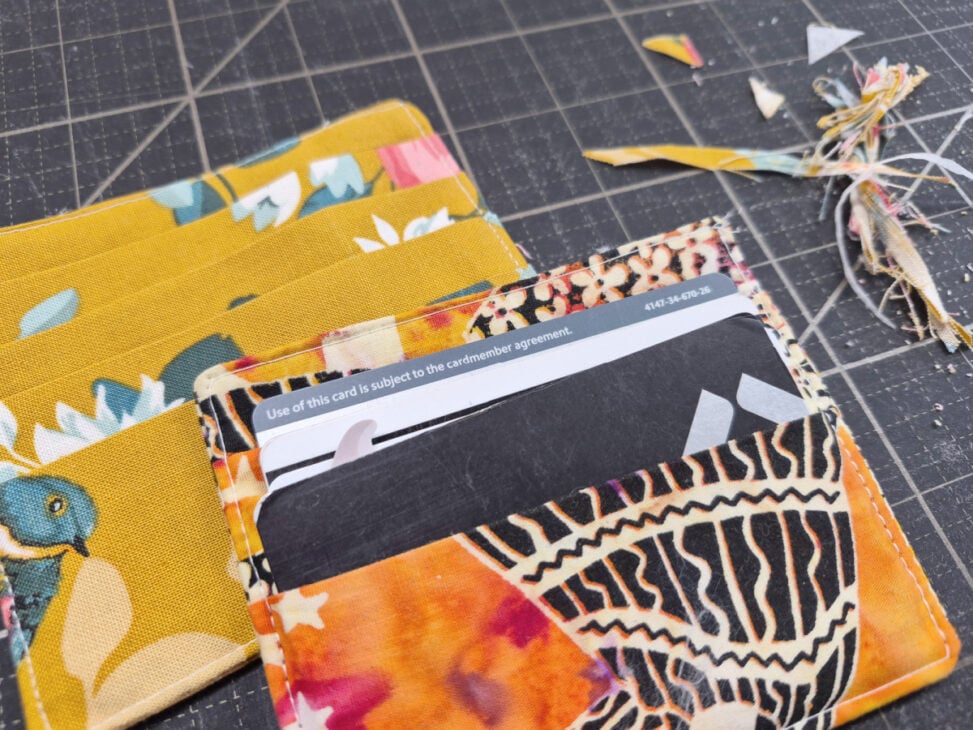 A handmade fabric wallet with colorful patterns holds a credit card. Scraps of fabric and thread are scattered on a black cutting mat, hinting at an easy to sew project for anyone looking for a simple card wallet pattern.