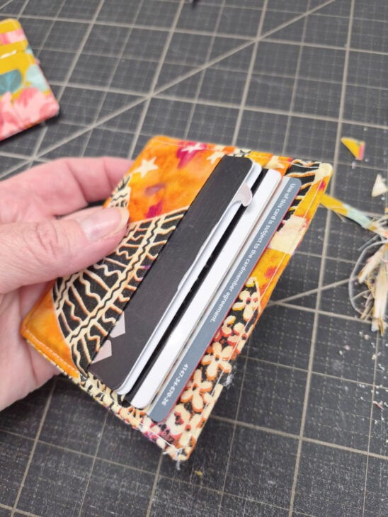 A hand holds an open fabric wallet with colorful patterns, revealing several credit cards inside. The wallet, made from an easy to sew card wallet pattern, rests on a black cutting mat with measurement lines and fabric scraps nearby.