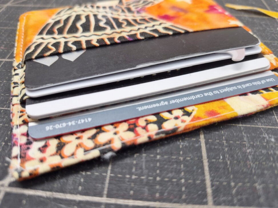 A colorful card wallet pattern lies open on a black grid surface, displaying three visible cards&mdash;including a partially exposed credit card&mdash;neatly tucked into its slots. This easy to sew design makes it both stylish and practical.