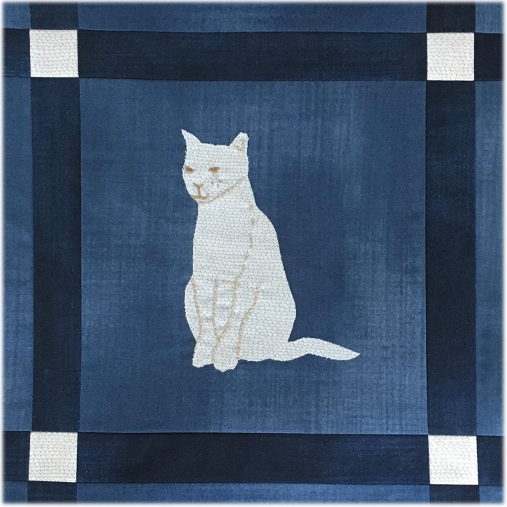A charming cat quilt block featuring an appliqu&eacute; of a white cat sitting on a blue background, bordered by a dark blue and white patchwork frame.