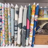 A shelf holds neatly organized rolls of colorful patterned fabric, a small tin with sailboat art, a yellow-lidded container, and a wooden box labeled "Larks EXTRA MILD" containing paper and items.