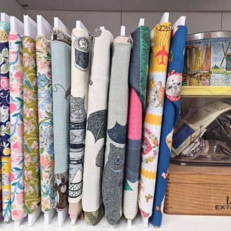 A shelf holds neatly organized rolls of colorful patterned fabric, a small tin with sailboat art, a yellow-lidded container, and a wooden box labeled "Larks EXTRA MILD" containing paper and items.